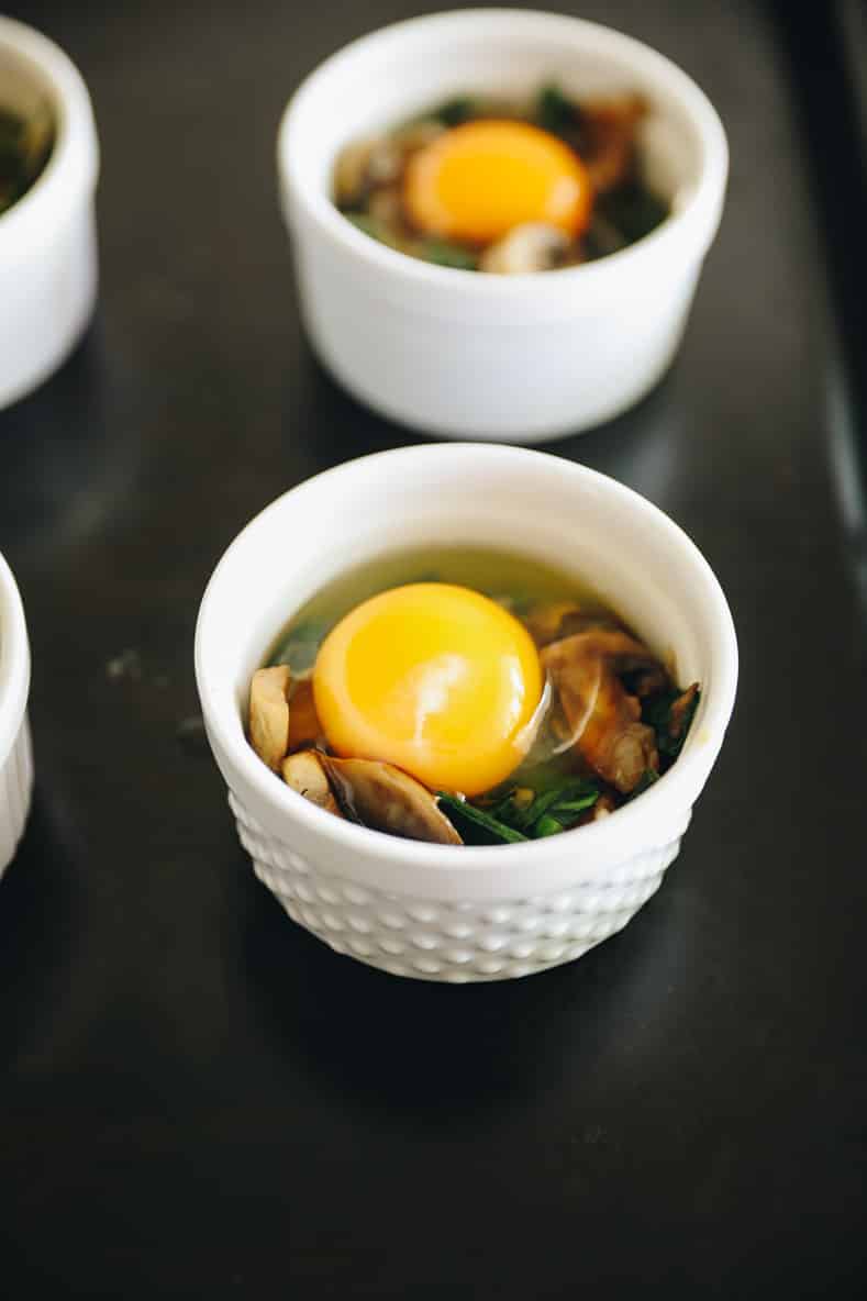 Two ramekins with vegetables and raw eggs cracked inside sitting on a baking sheet.