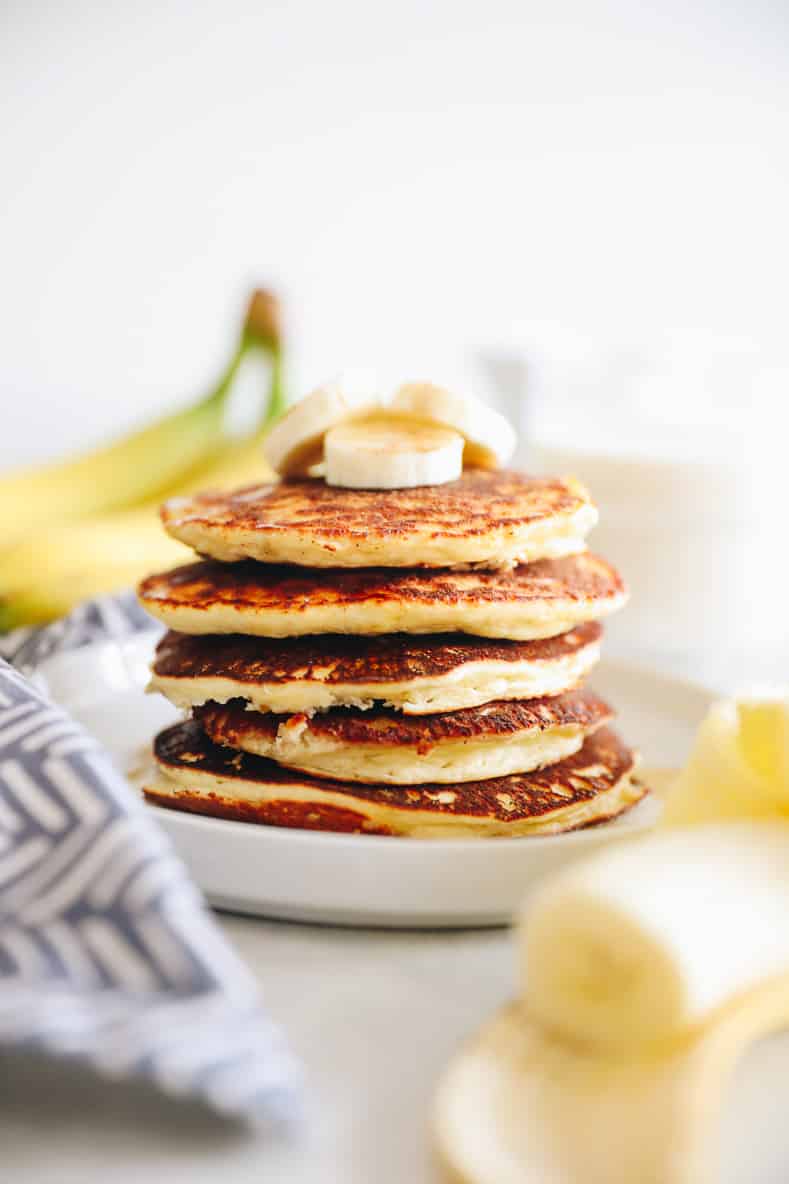 Banana protein pancakes stacked on a white plate. Bananas in foreground and background of the image.