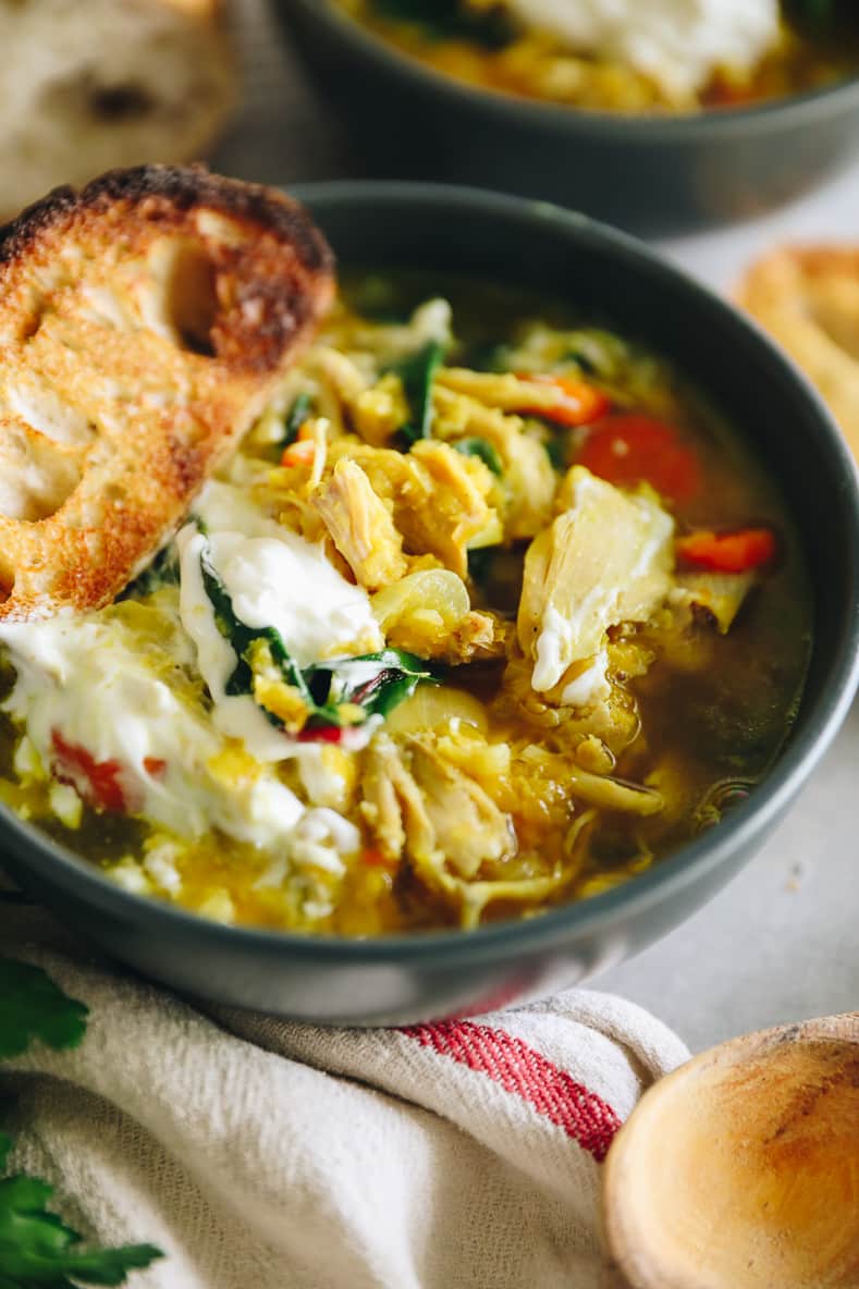 Angled shot of chicken lentil soup in a dark grey bowl. Soup is topped with sour cream and a piece of crunchy bread.