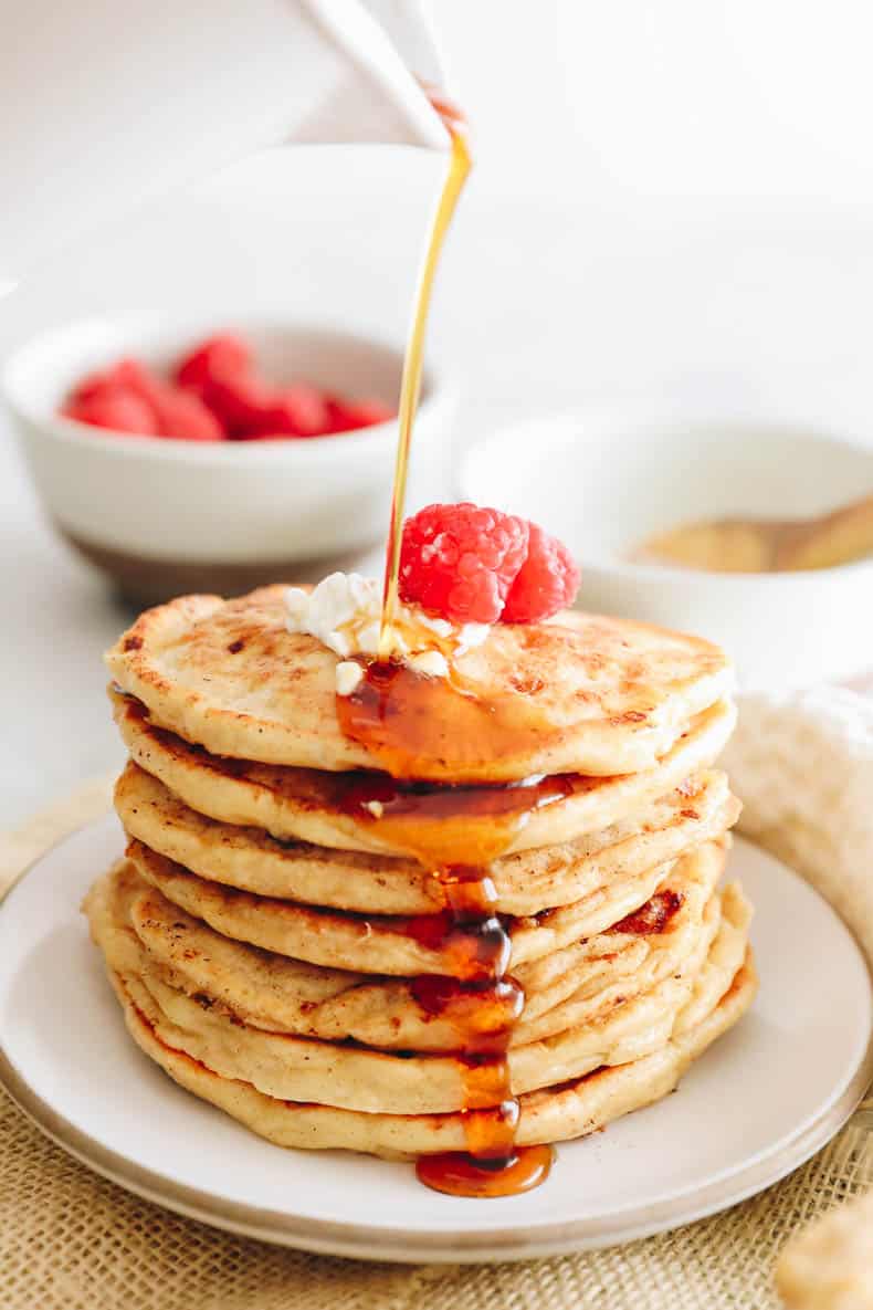 maple syrup being poured on a stack of pancakes with raspberries and cottage cheese.