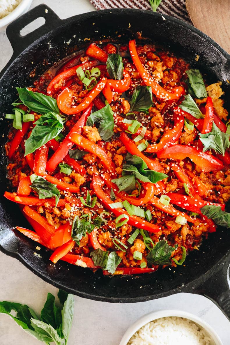 overhead image of gochujang chicken cooked in a skillet with veggies, basil, sesame seeds and green onions.