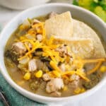 Green chili in a white bowl with tortilla chips and shredded cheddar cheese.