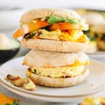 Two meal prep breakfast sandwiches inside english muffins on a white plate with avocado, tomato and cheese.