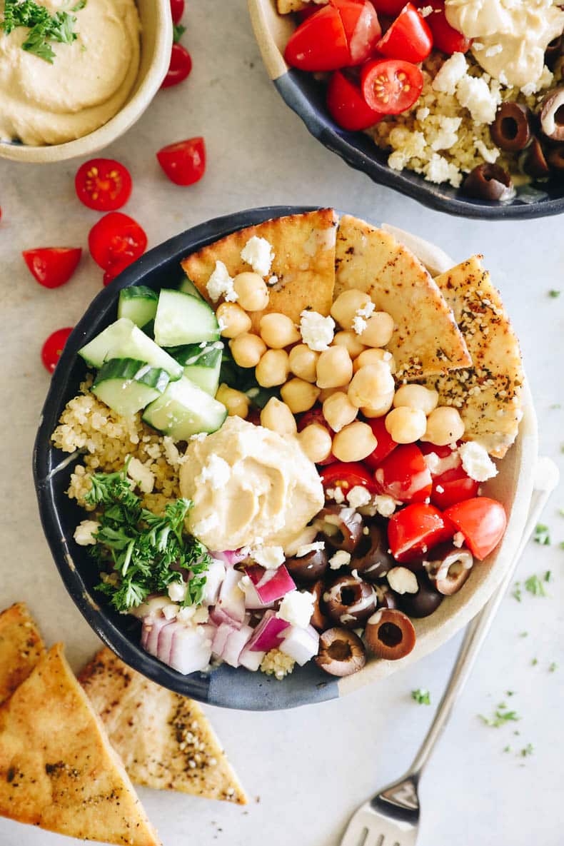 overhead photo of a mediterranean bowl with quinoa, olives, feta, pita chips, hummus, tomatoes and red onion.