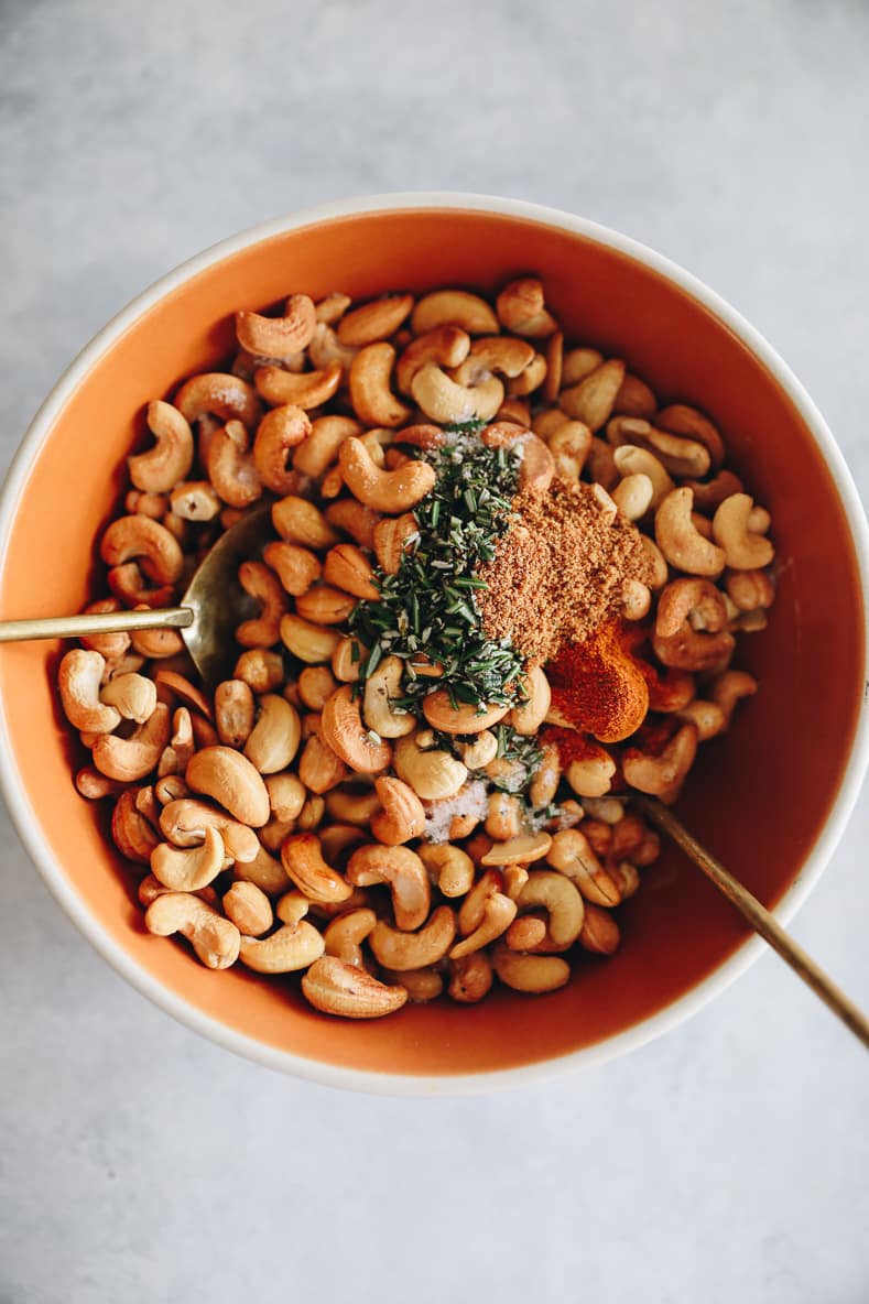 An orange bowl with cashews, rosemary and additional spices in it. Two spoons are placed in the bowl for mixing.