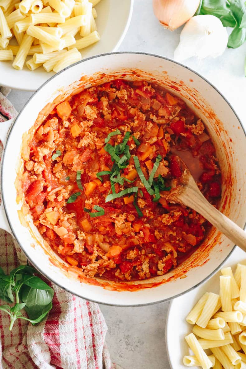 cooked turkey bolognese with plain rigatoni noodles in a white dutch oven with a wooden spoon.