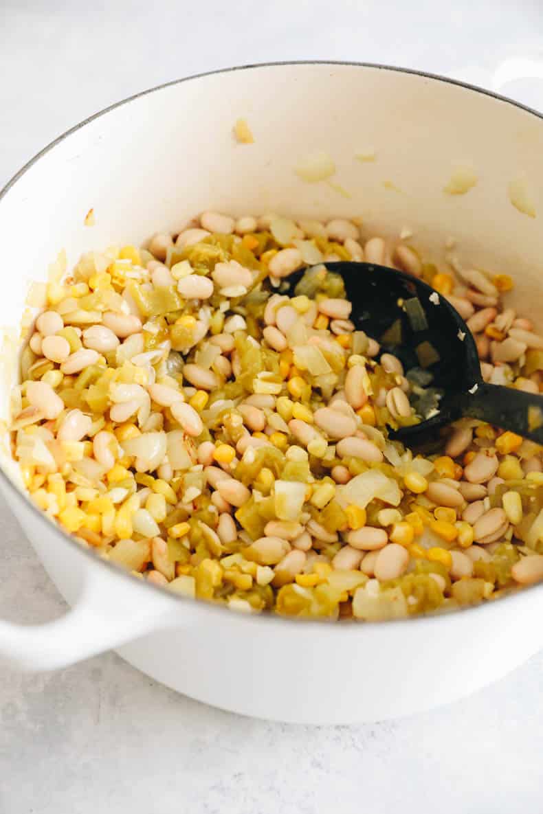 onions, garlic, white beans, corn and green chiles in a white dutch oven.